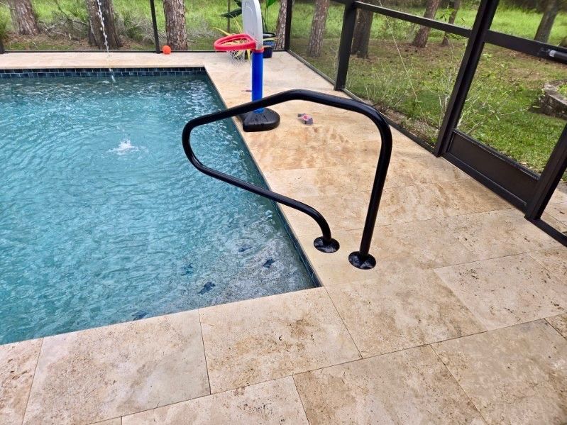 Pool with black handrail, surrounded by beige tile, screened enclosure, and trees.