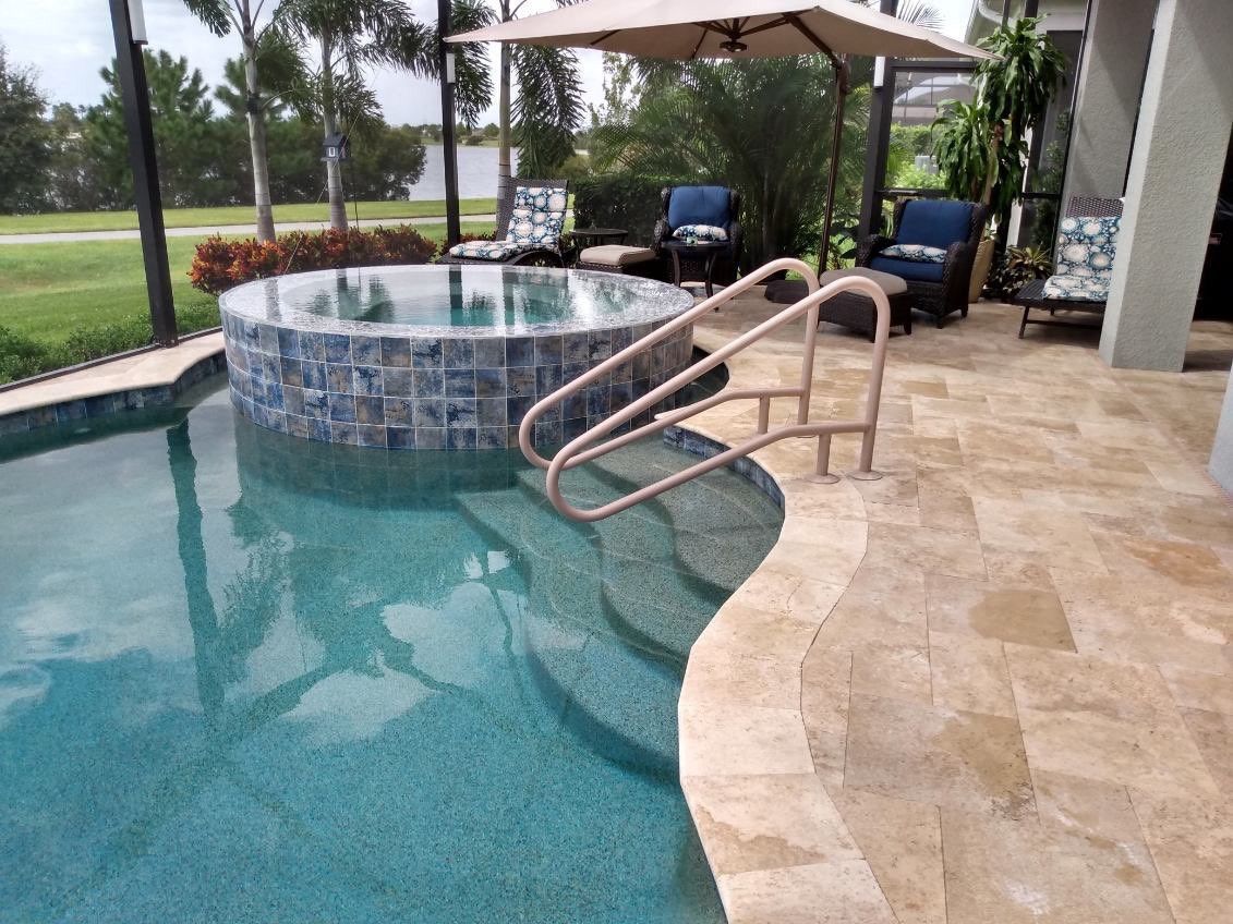 Pool with spa, steps, handrail. Beige patio, blue tile spa. Chairs, umbrella, green lawn in background.