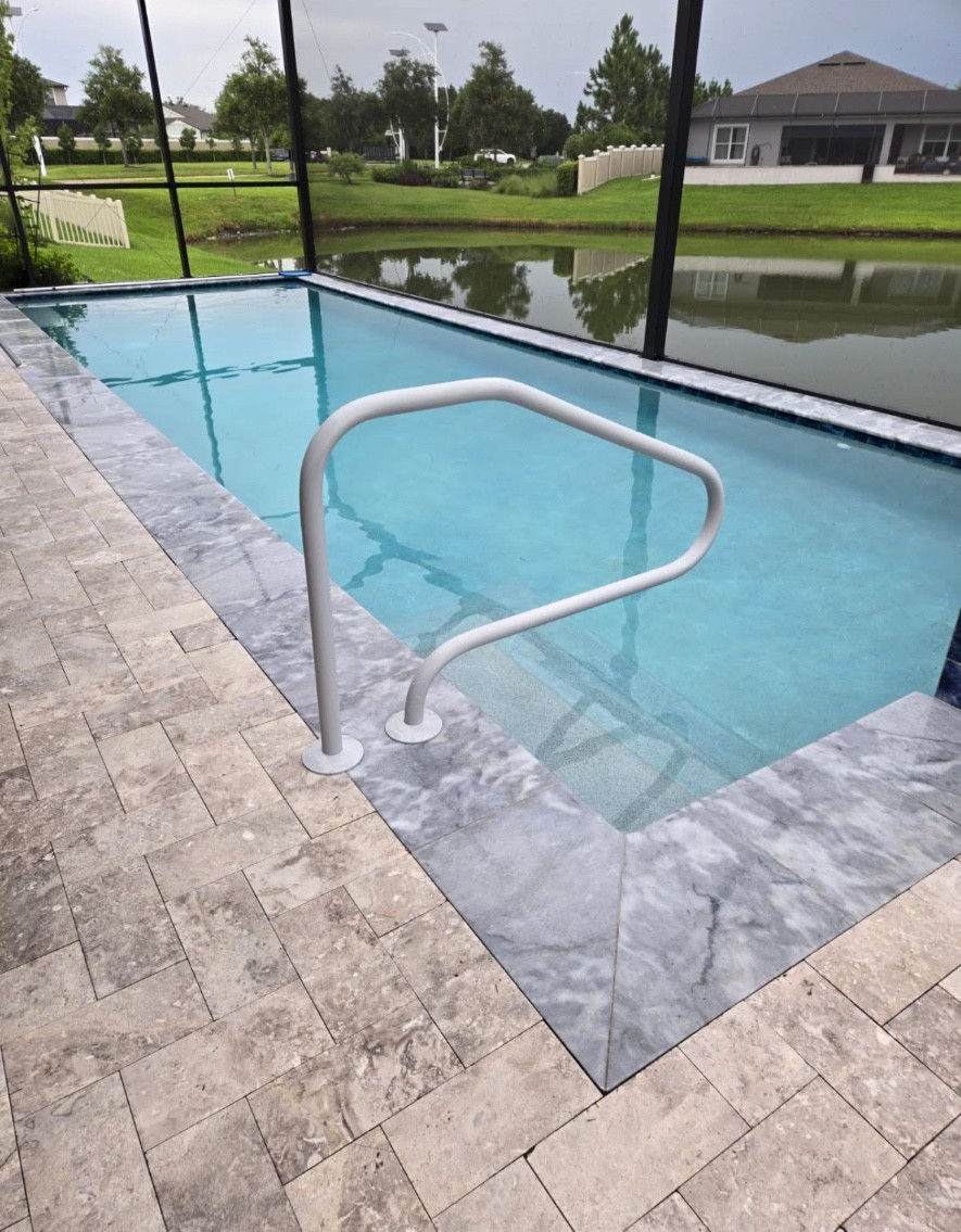 Small rectangular pool with gray tile, handrail, and surrounding stone pavers. In the background, houses and a pond are visible.