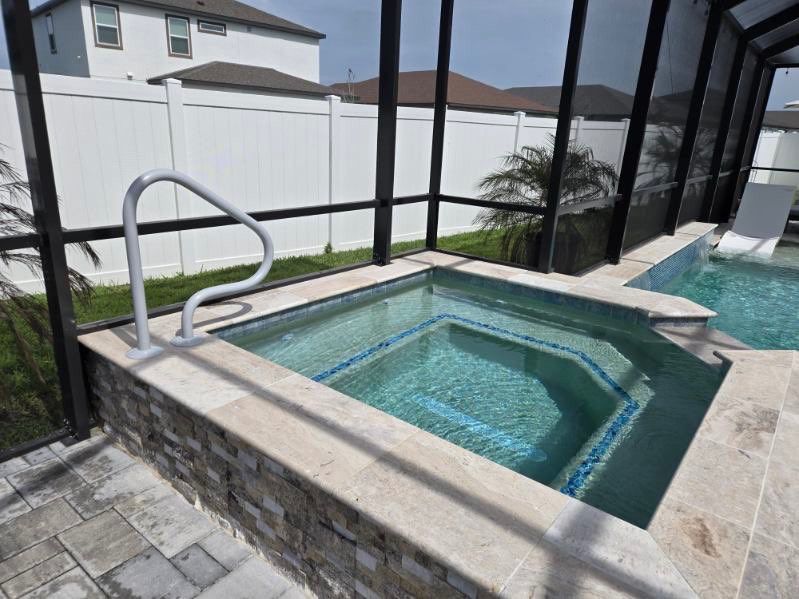 Hot tub with light stone surround, connected to a pool, enclosed by a screened patio.