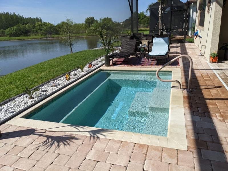 Small turquoise pool with stairs next to a lake, surrounded by brick patio and beige trim.