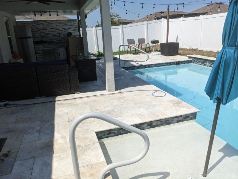 Poolside patio with a pool, white fence, and seating area. Blue umbrella in the foreground.