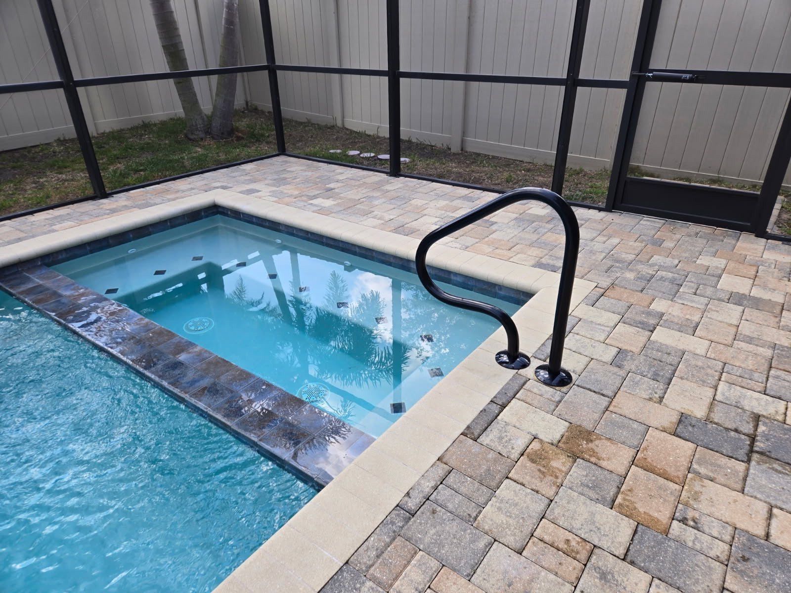 Pool with spa, brick patio, black handrail. Fenced yard with a door.