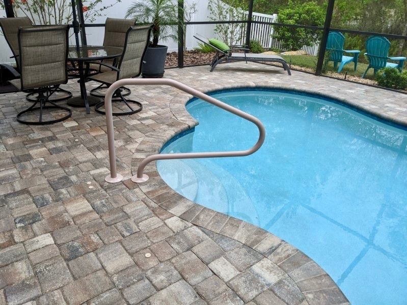 A pool with a tan brick patio, a metal handrail, and outdoor furniture.