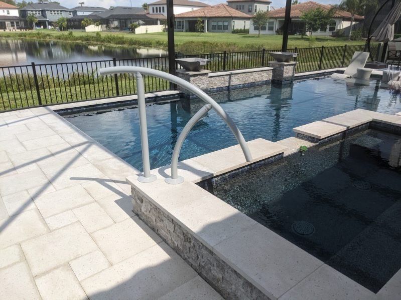Swimming pool with blue water, gray handrail, and light stone patio, with a pond and houses in the background.