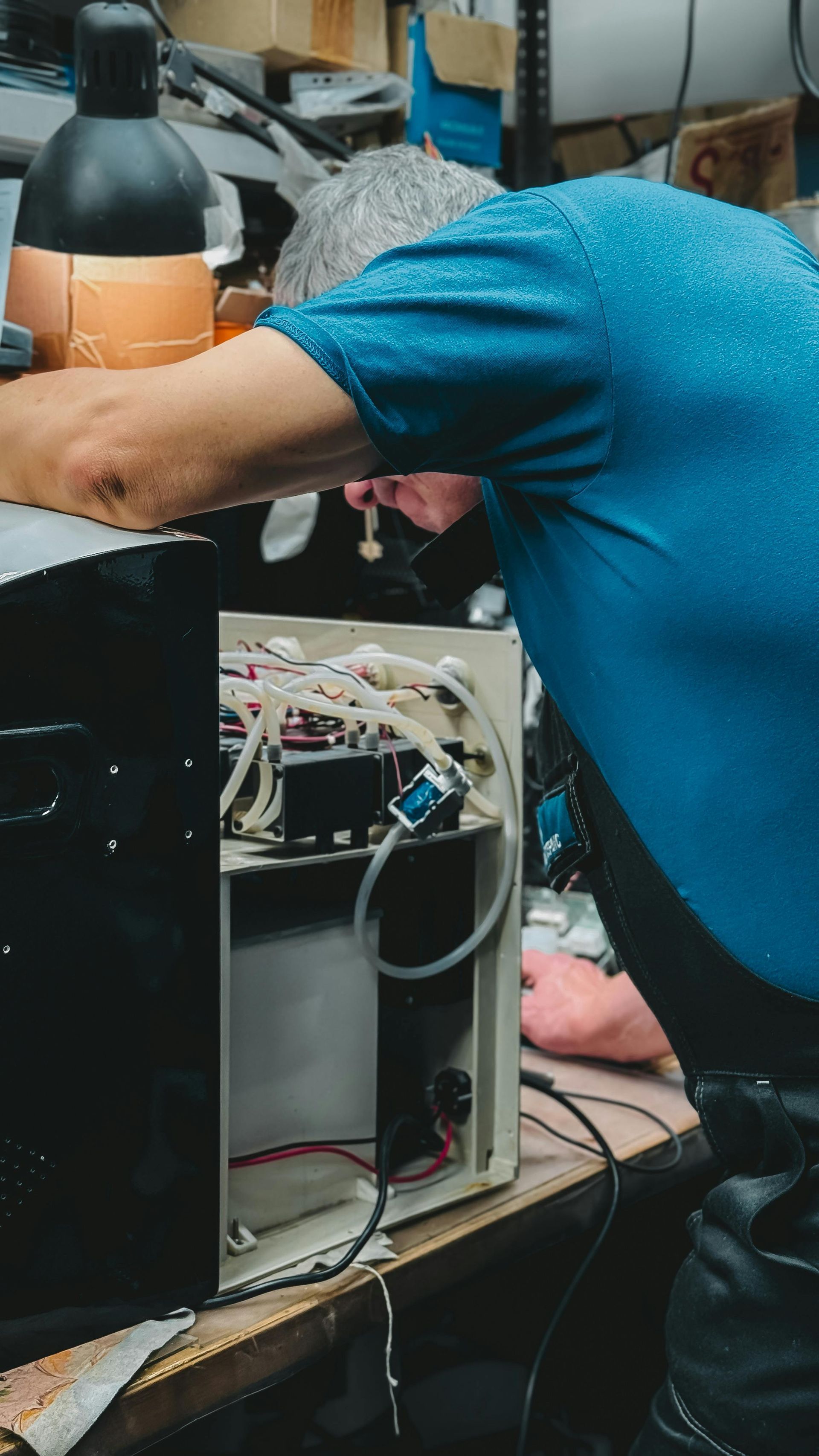 Man in blue shirt repairing a machine, with open panel revealing internal components. Workshop setting.