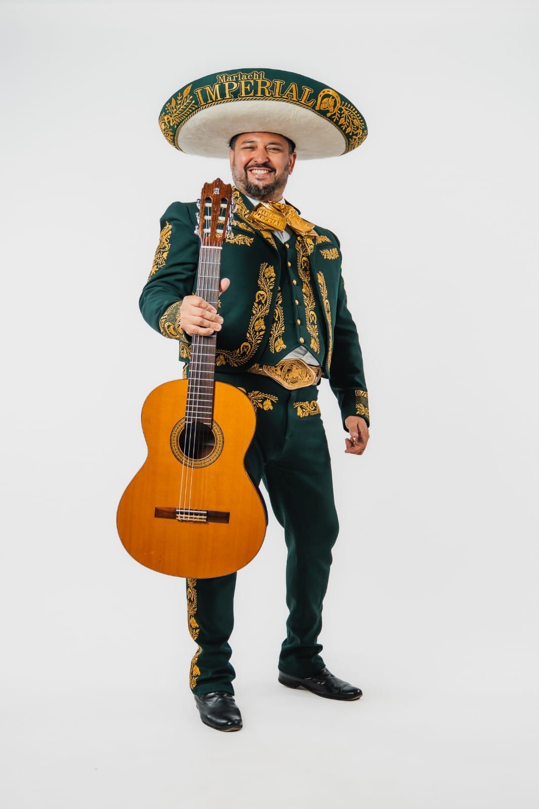 Un músico sonriente con un traje de mariachi verde oscuro y un sombrero, sosteniendo una guitarra