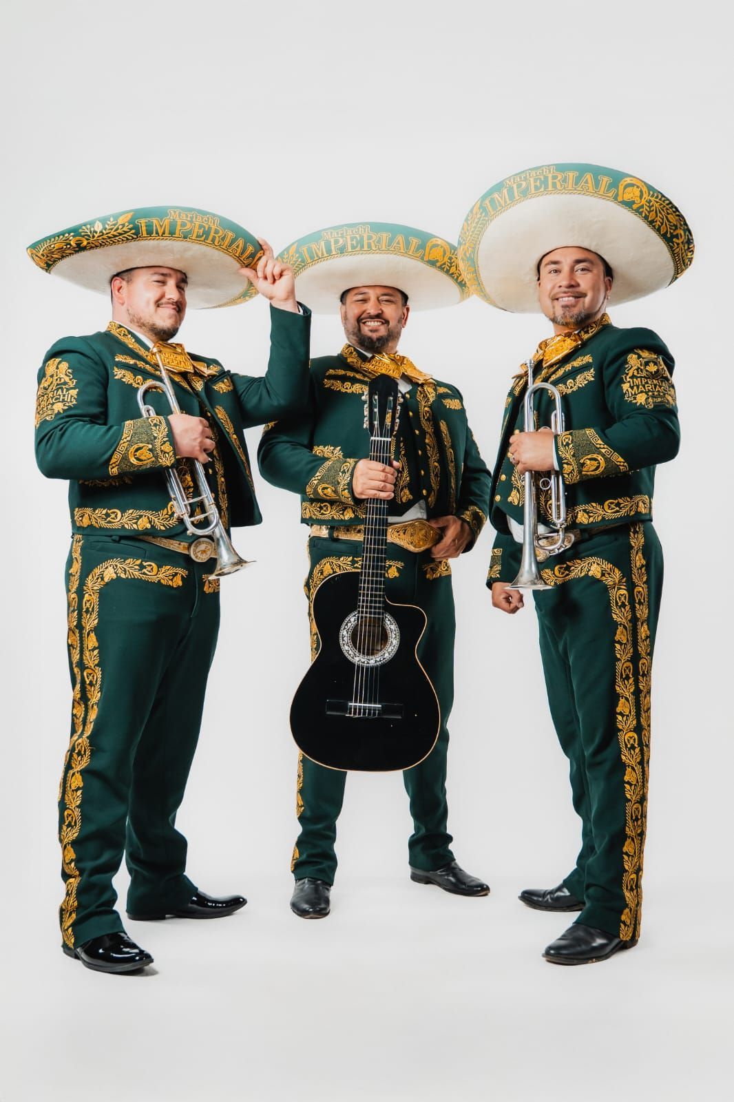Tres músicos con trajes de mariachi verde oscuro con bordados dorados y sombreros, de pie sobre un fondo blanco.