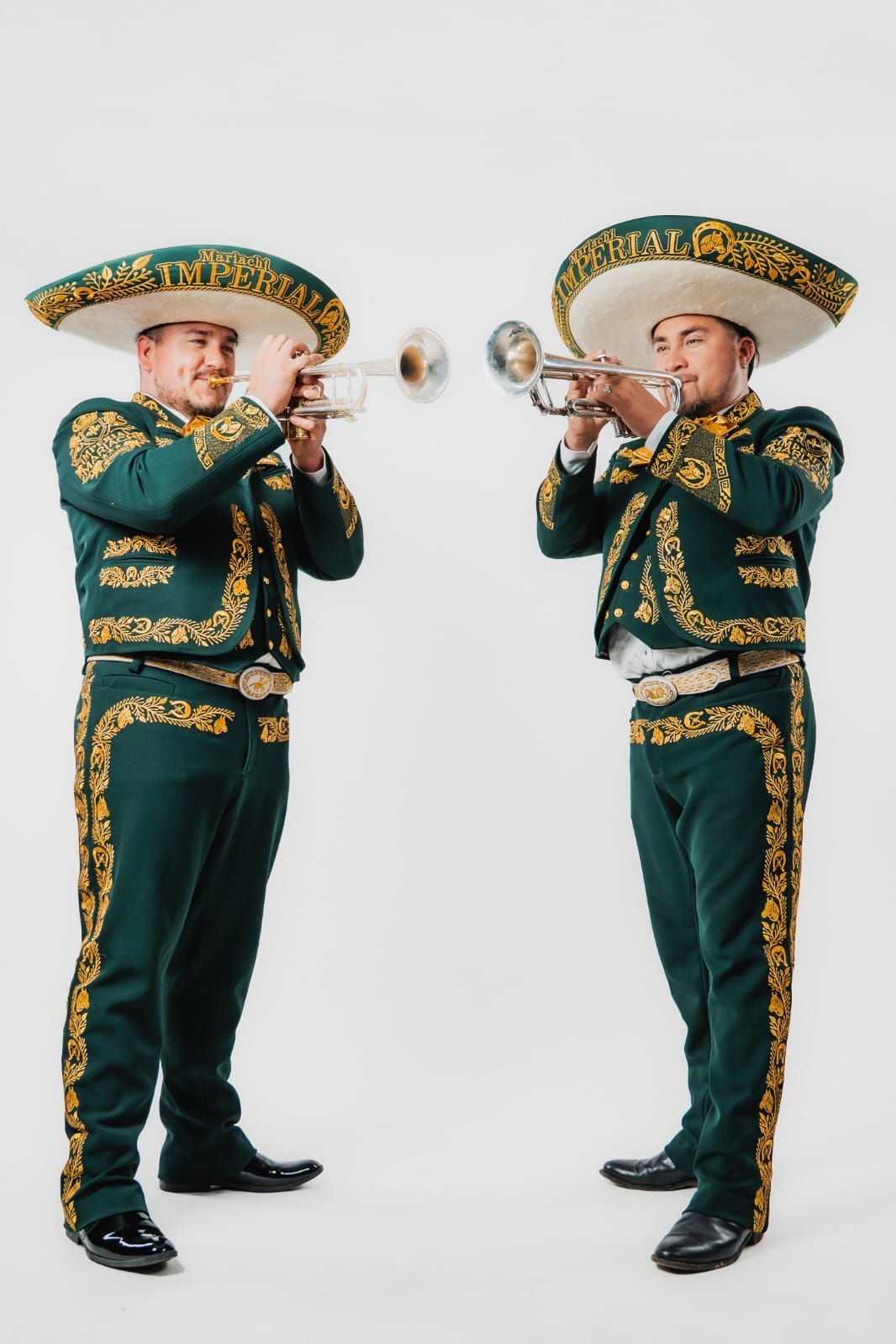 Dos músicos de mariachi con trajes verdes y dorados y sombreros a juego tocan las trompetas sobre un fondo blanco.