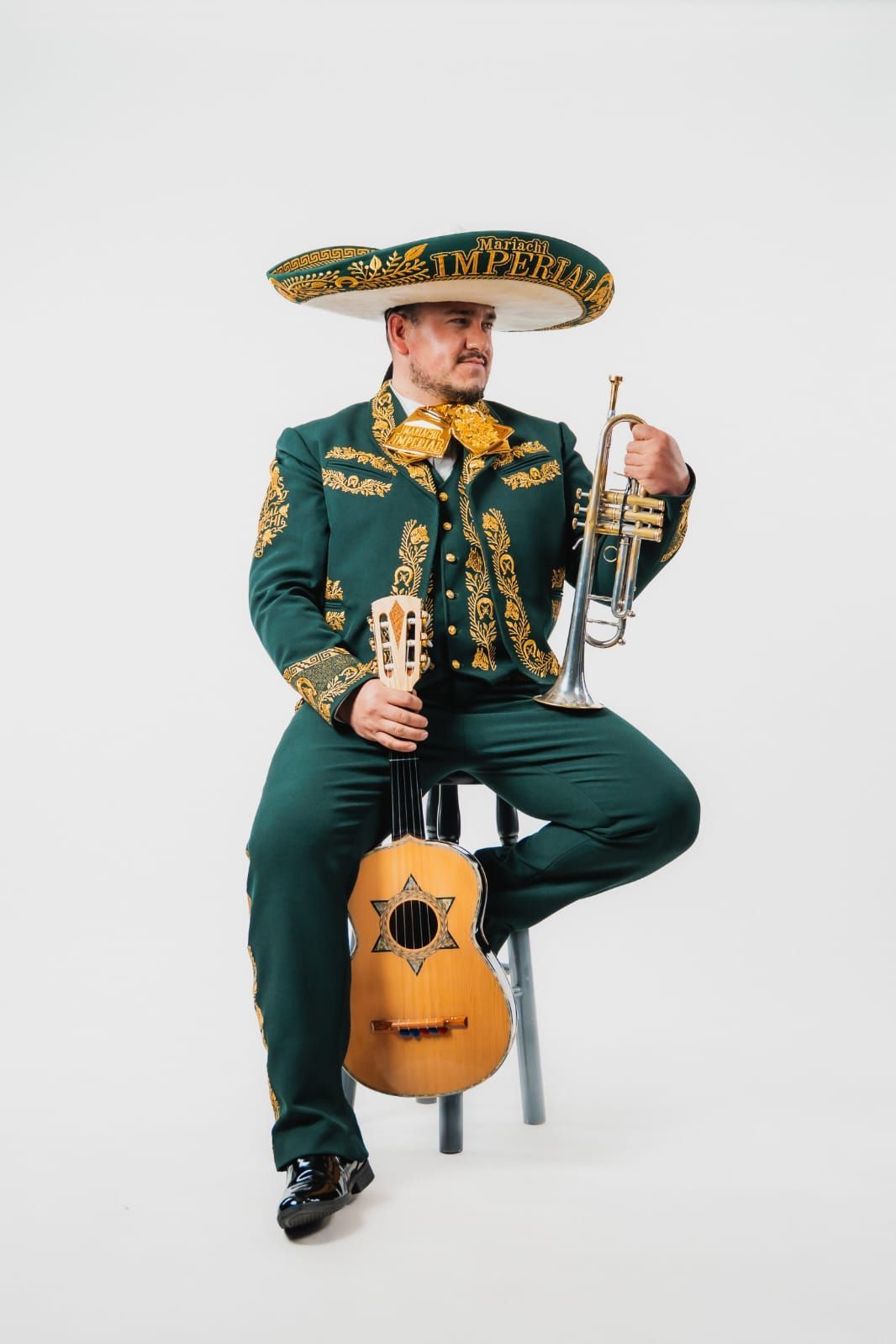 Un músico con un traje de mariachi verde y dorado está sentado en un taburete, sosteniendo una trompeta