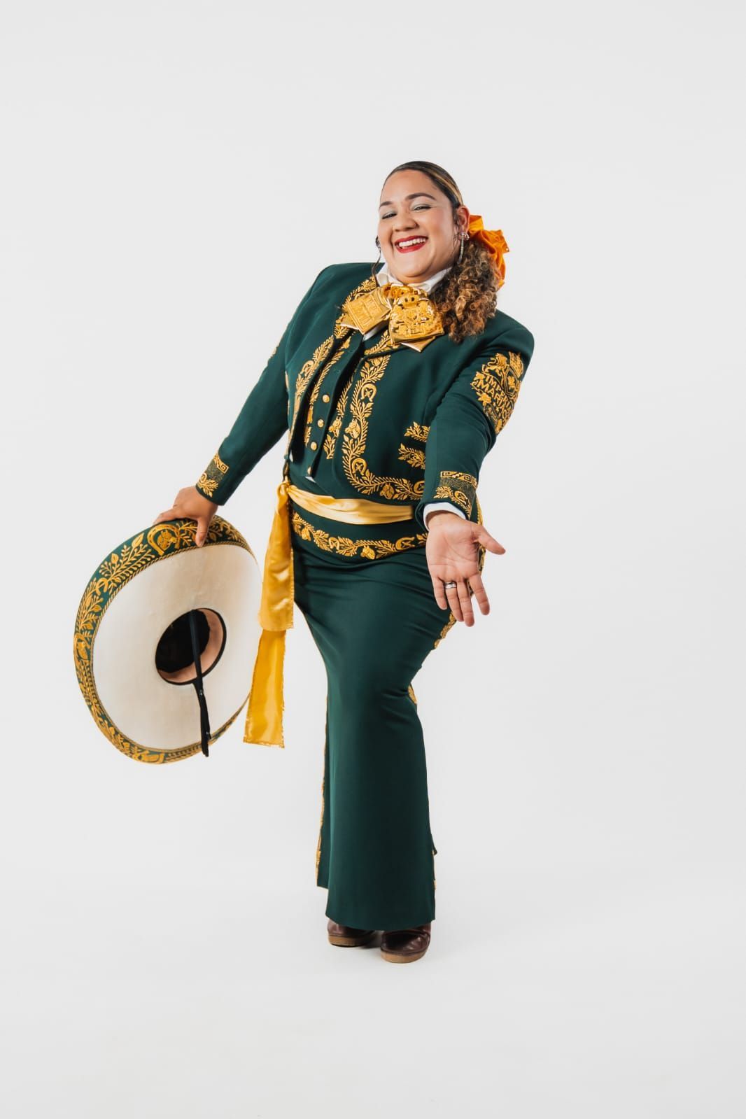Un artista sonriente, vestido con un traje de mariachi verde oscuro con bordados dorados, sostiene un sombrero