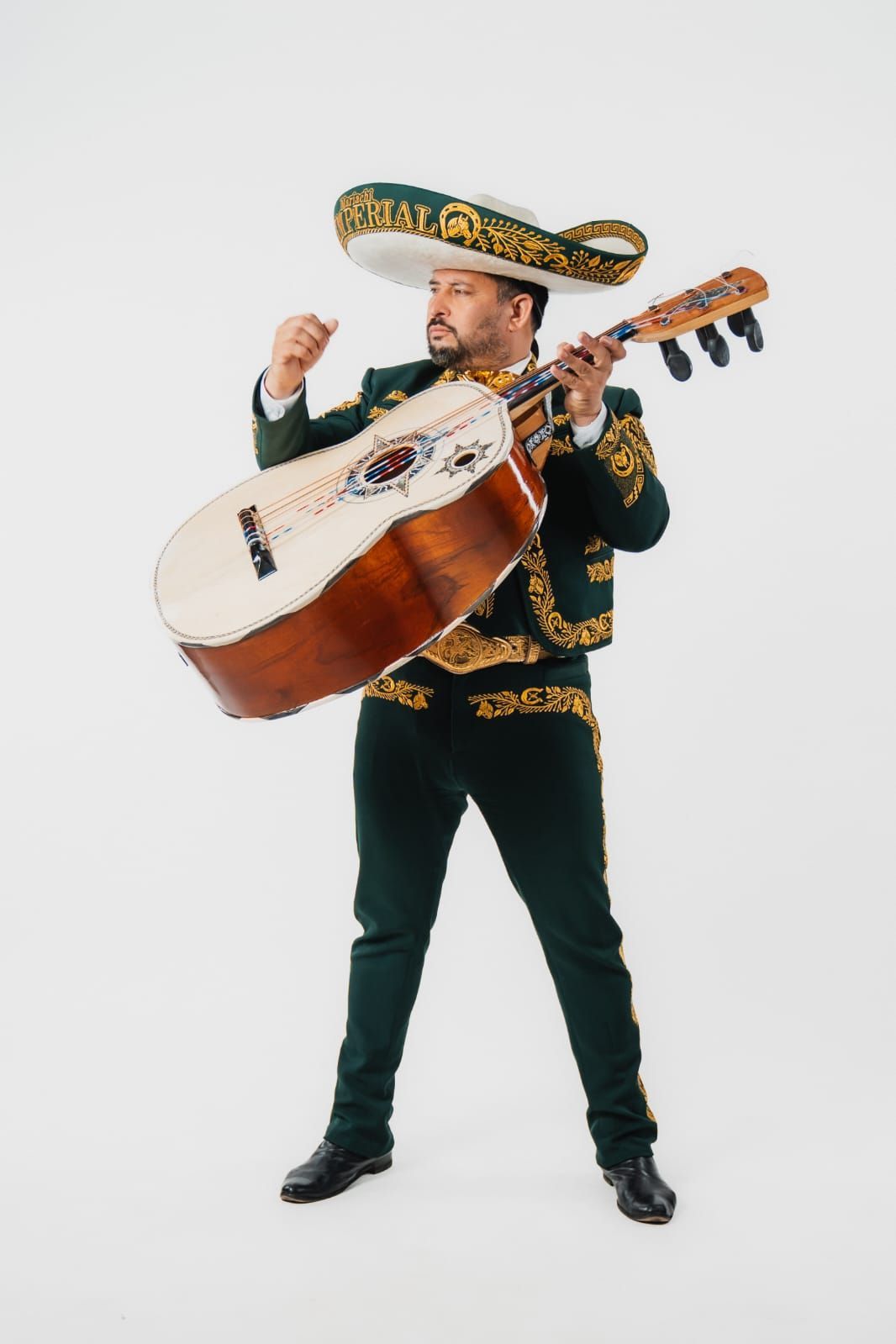 Un músico con traje de mariachi verde y sombrero tocando un guitarrón sobre un fondo blanco liso.