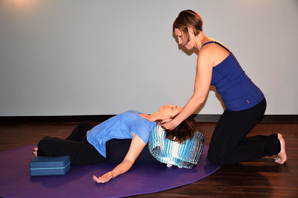Woman receiving neck massage, lying on mat with support, another woman kneels nearby.