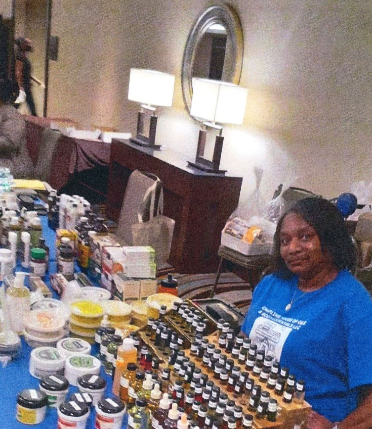 Woman at a table with products, possibly at a convention. The setting is indoors with lamps and a mirror.