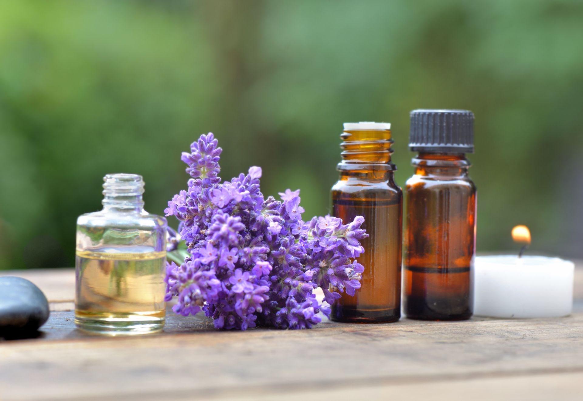 Bottles of essential oils, lavender flowers, lit candle, wooden surface, green background.