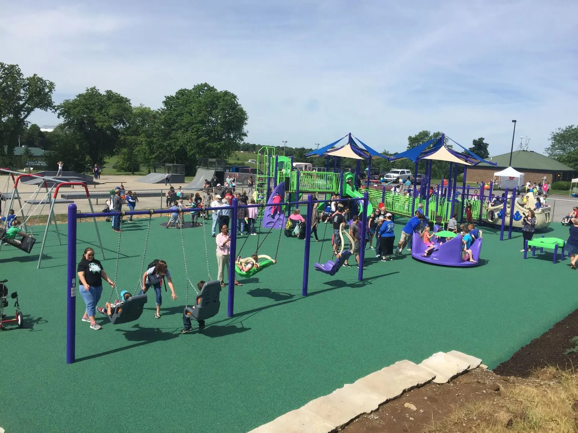 Children playing on a green playground with swings, slides, and other equipment under a sunny sky.