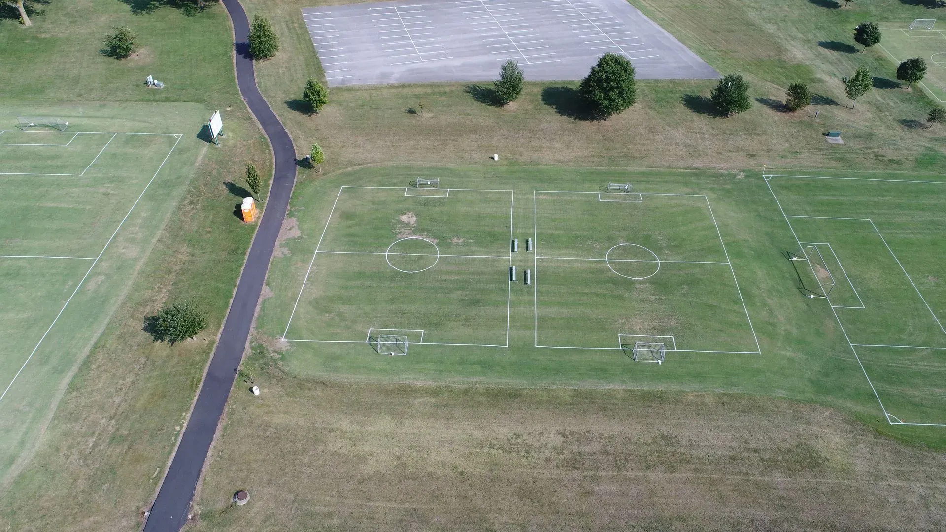 Aerial view of a green soccer field with white lines, asphalt path, and trees.