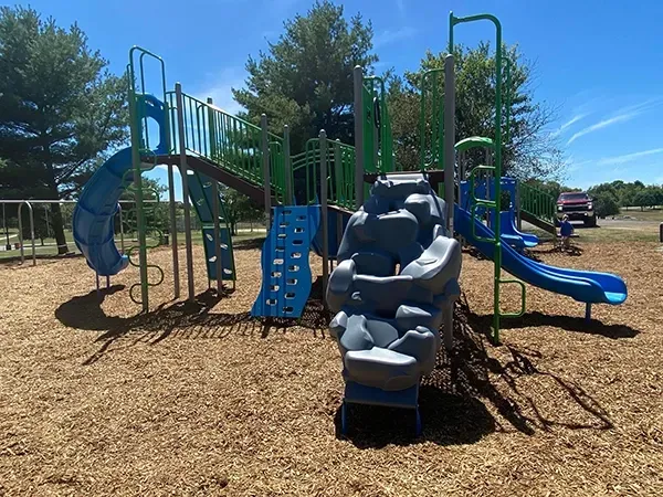 Playground with blue and green structures on wood chips, under a blue sky.