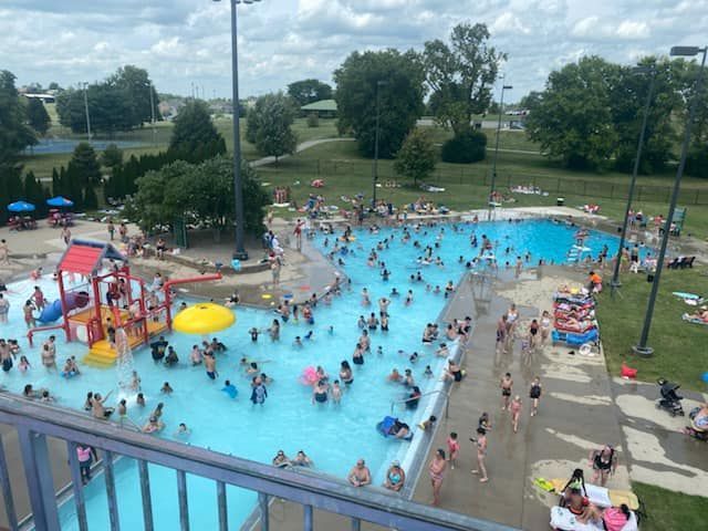Crowded outdoor pool with people swimming, a water slide, and green park setting.