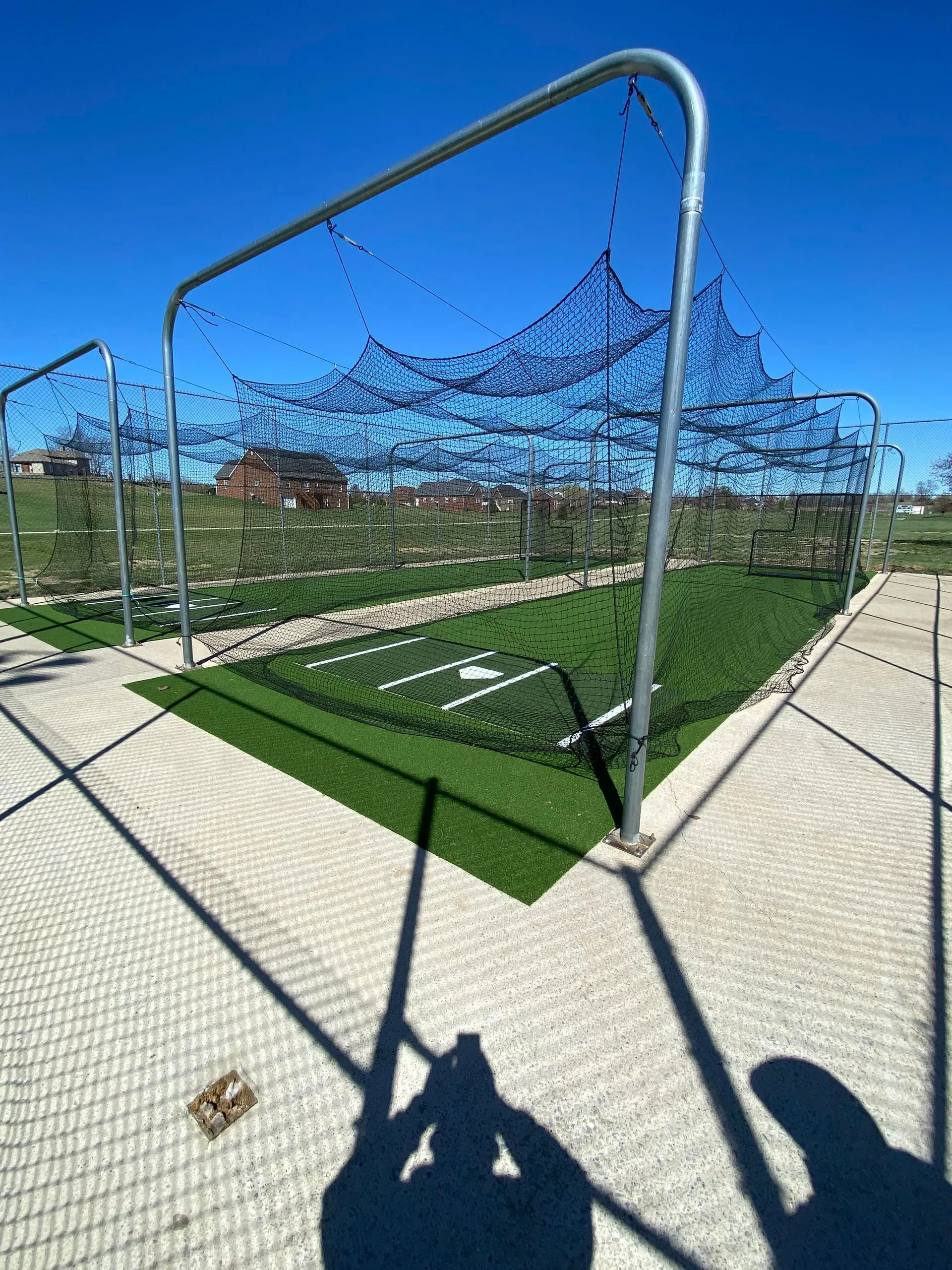A baseball batting cage on artificial turf under a bright blue sky. A person's shadow is cast in the foreground.