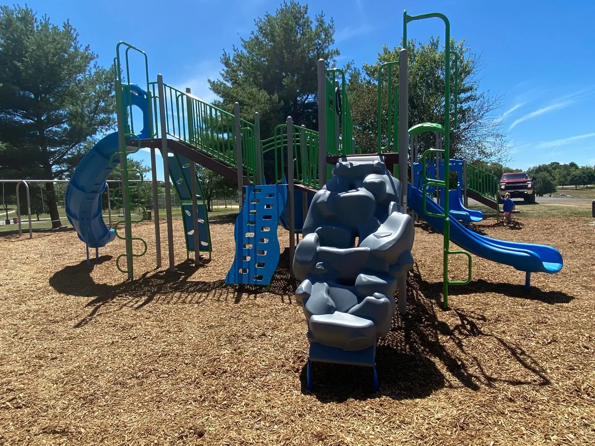 Playground with blue slides, climbing walls, and a rock-climbing feature set on wood chips, under a sunny sky.