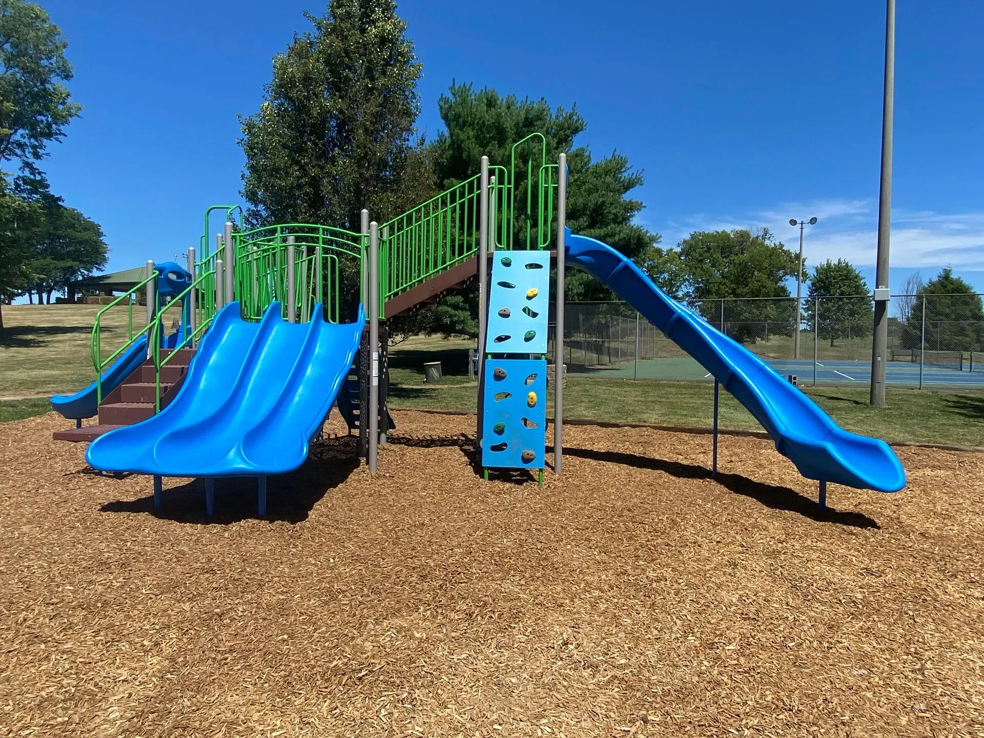 Blue and green playground with slides and climbing wall, set on wood chips, under a blue sky.