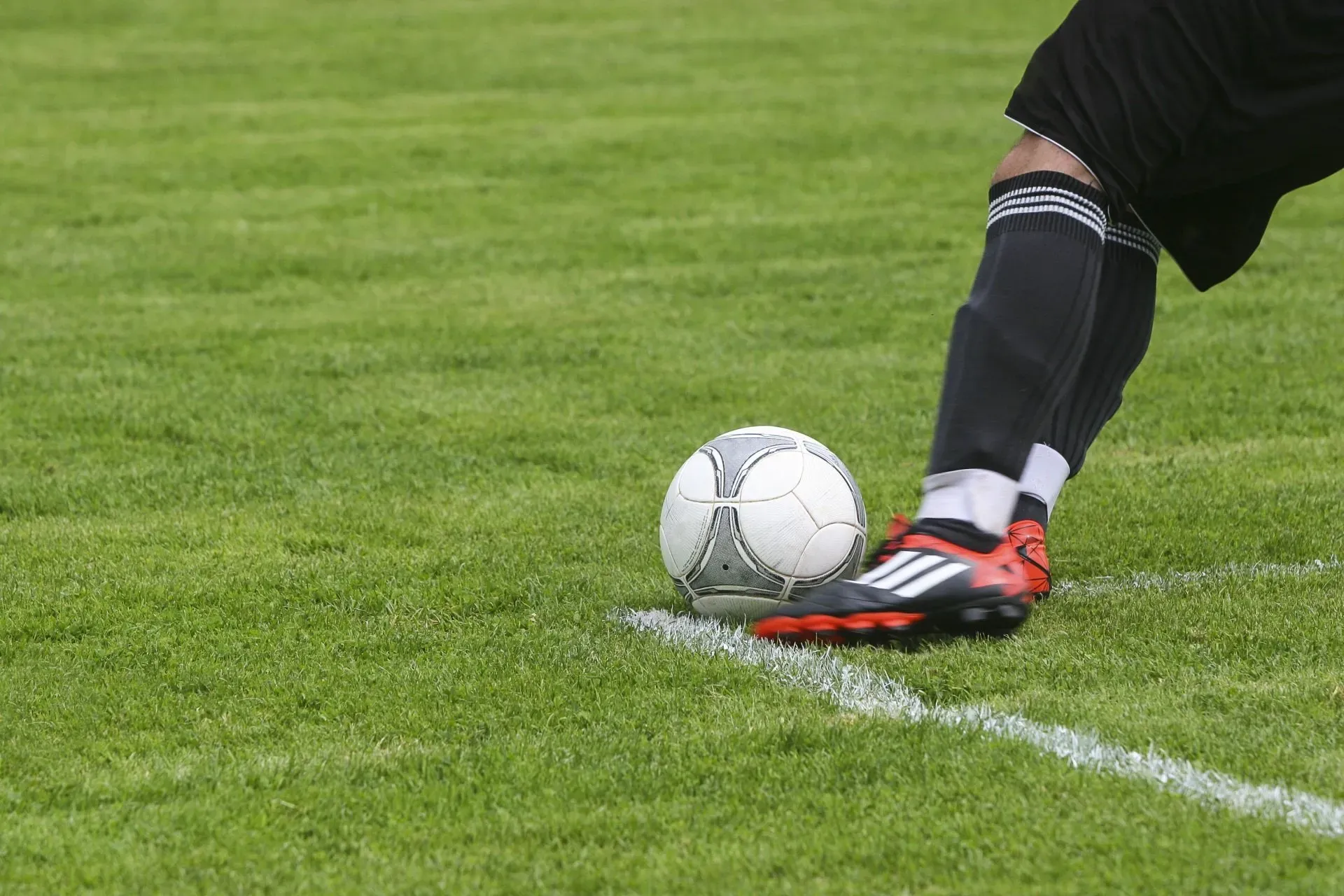 Soccer player kicking the ball at the edge of the field, grass field, black shorts and socks, red cleats.