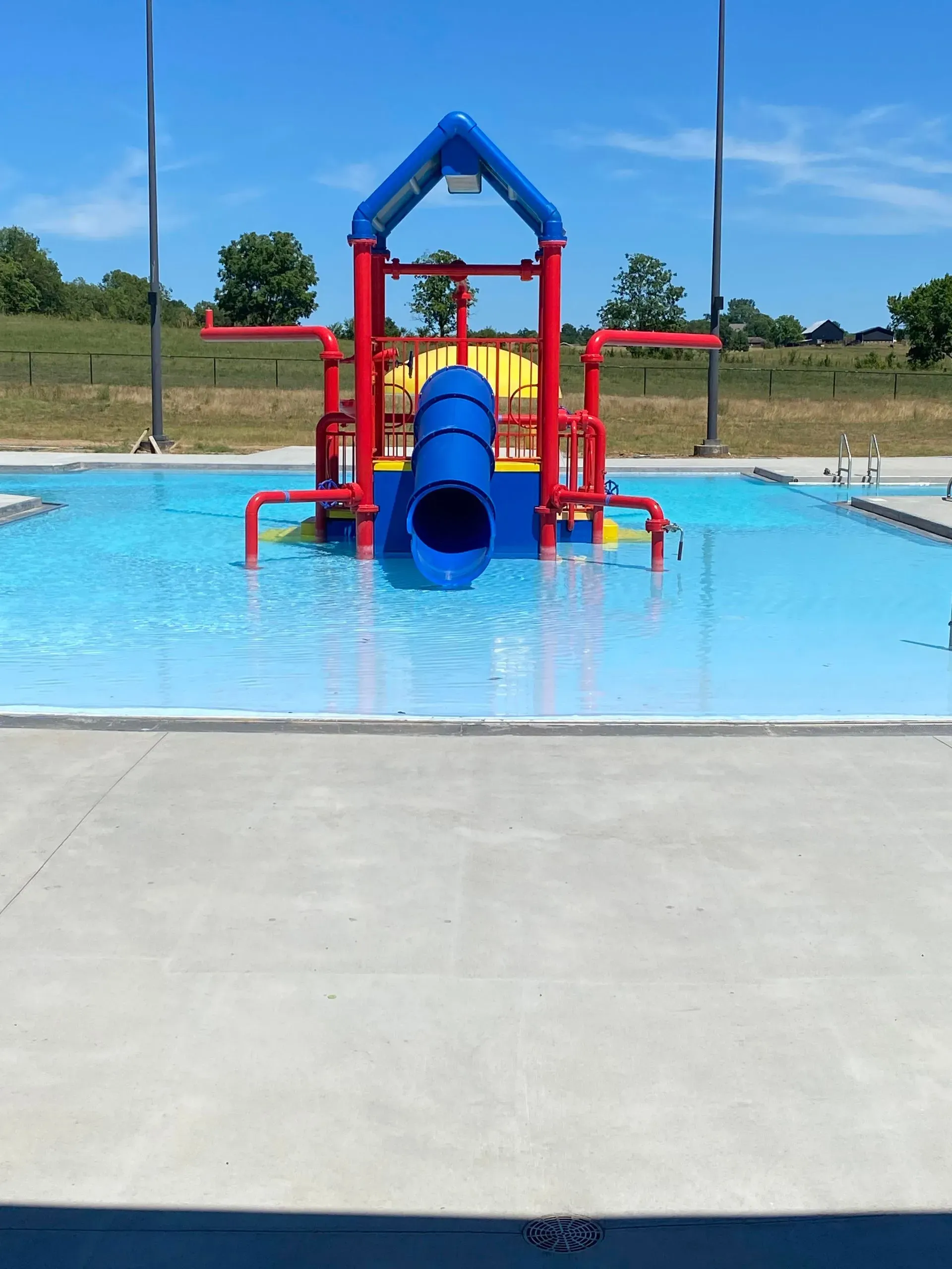 Swimming pool with blue slide, red and yellow structures, clear water, sunny day.