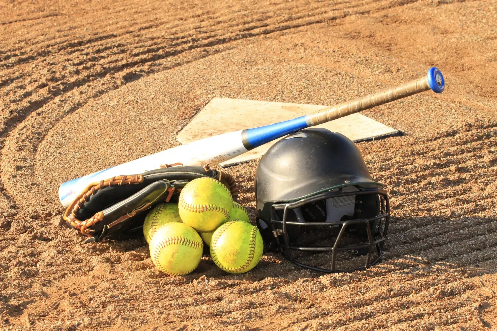 Softball gear on a dirt field: bat, helmet, glove, and yellow softballs near home plate.