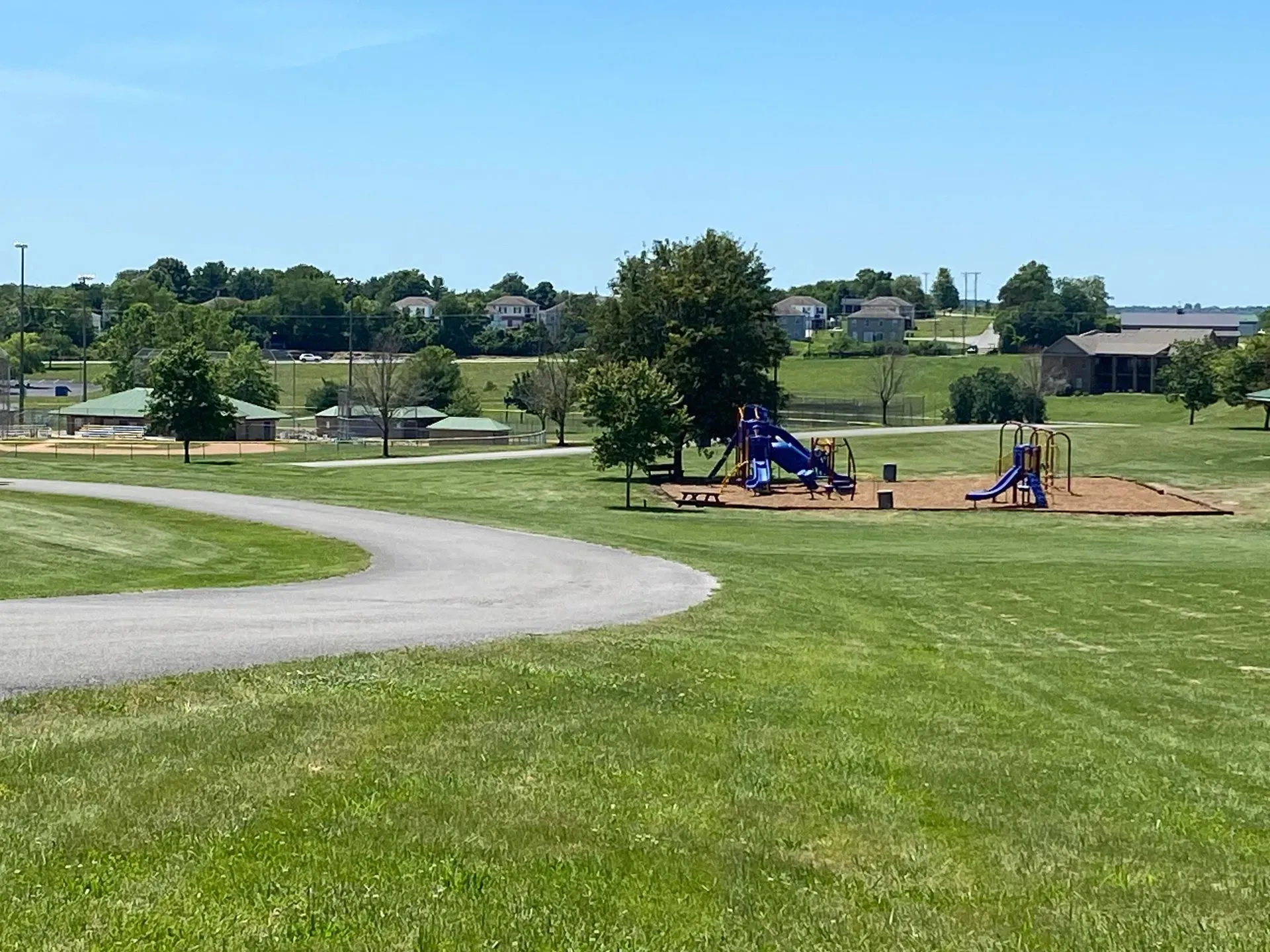 Park with a playground, walking path, and buildings under a blue sky.