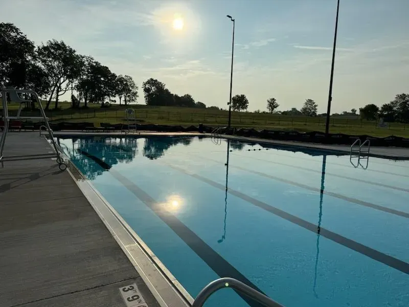 A swimming pool reflecting the sun, with lane markers, a concrete deck, and grassy hills in the background.