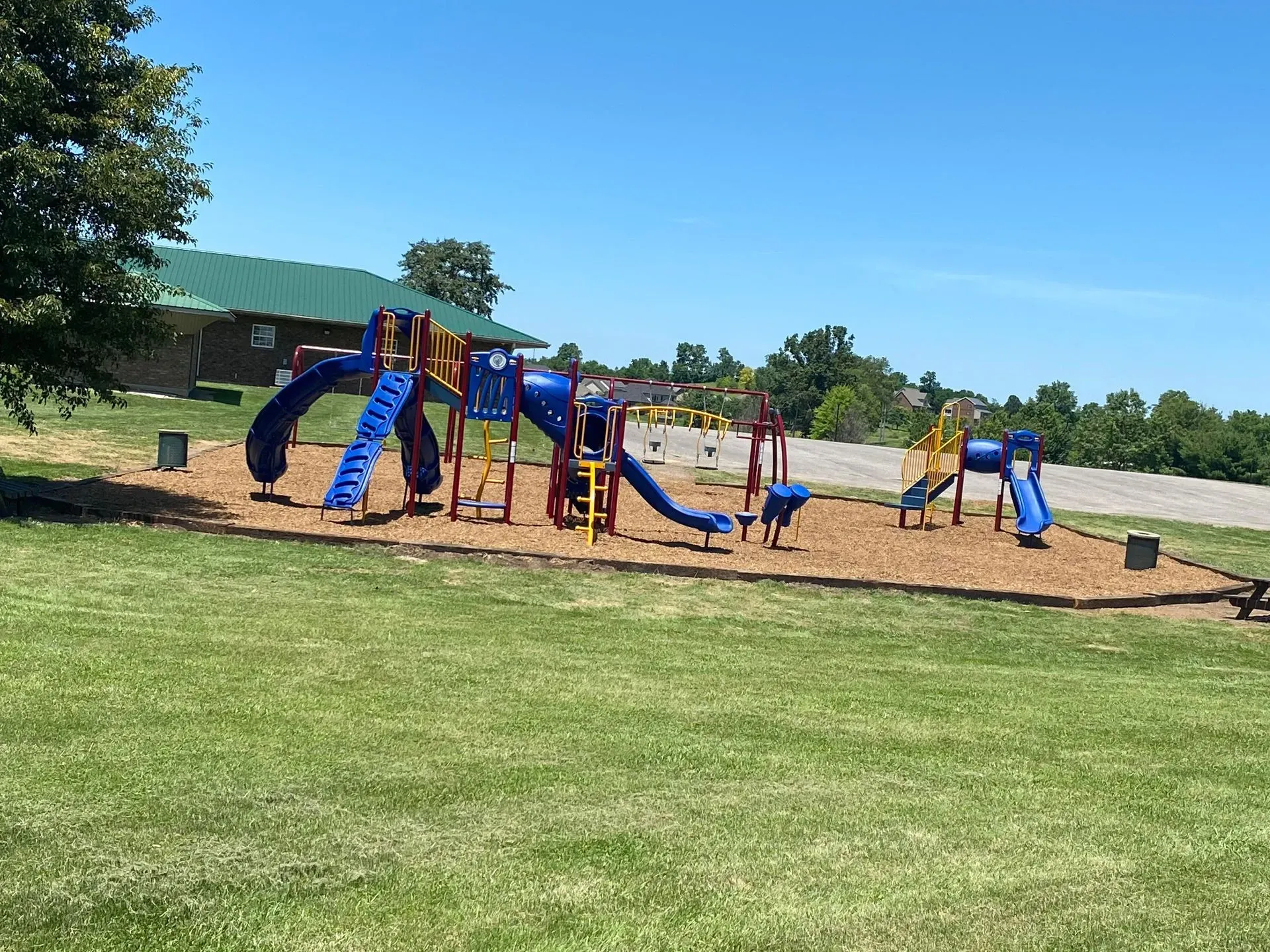 Playground with blue slides and red accents on wood chips, green grass, and a clear sky.