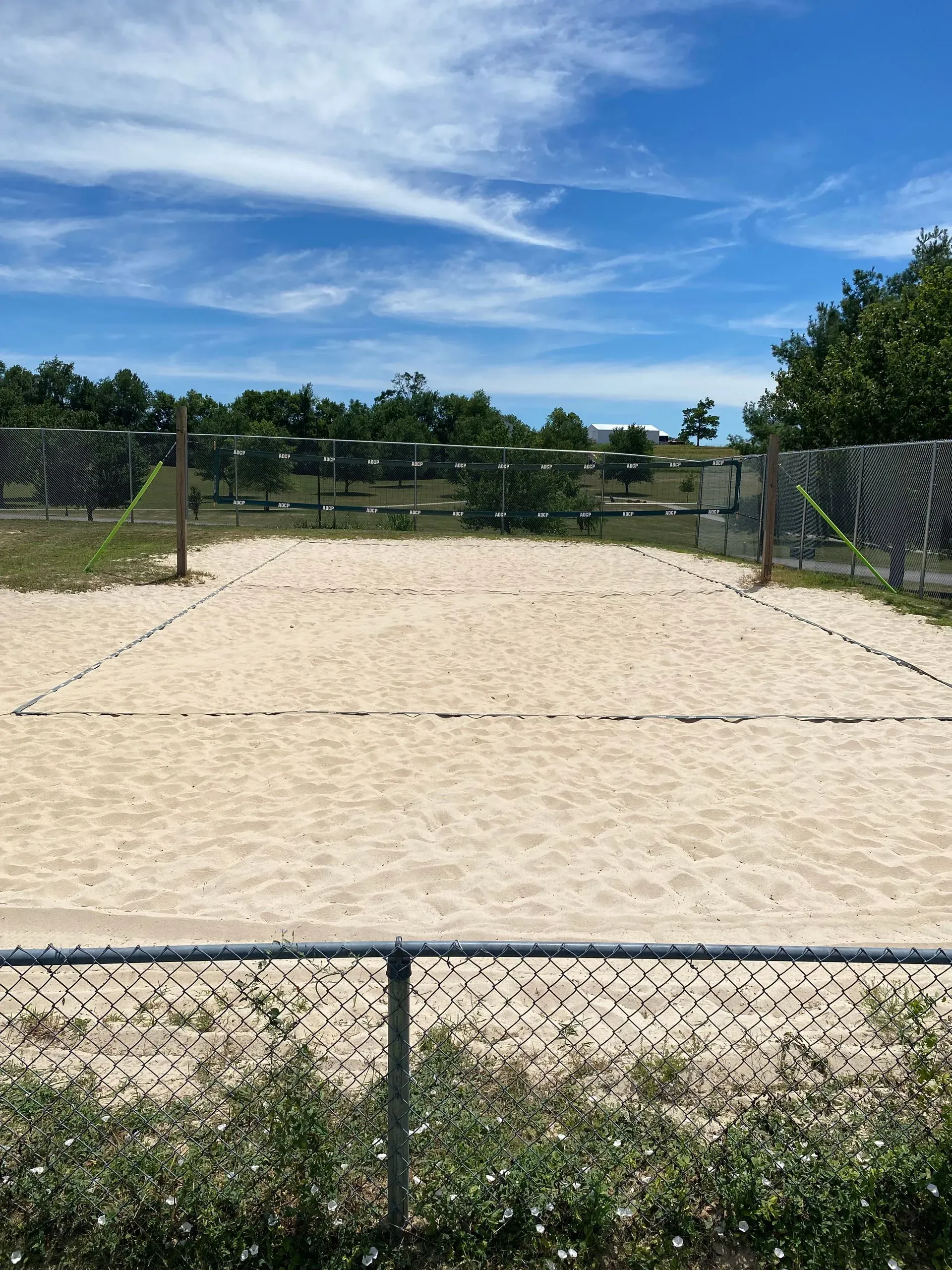 Empty sand volleyball court under a blue sky, viewed through a chain link fence.