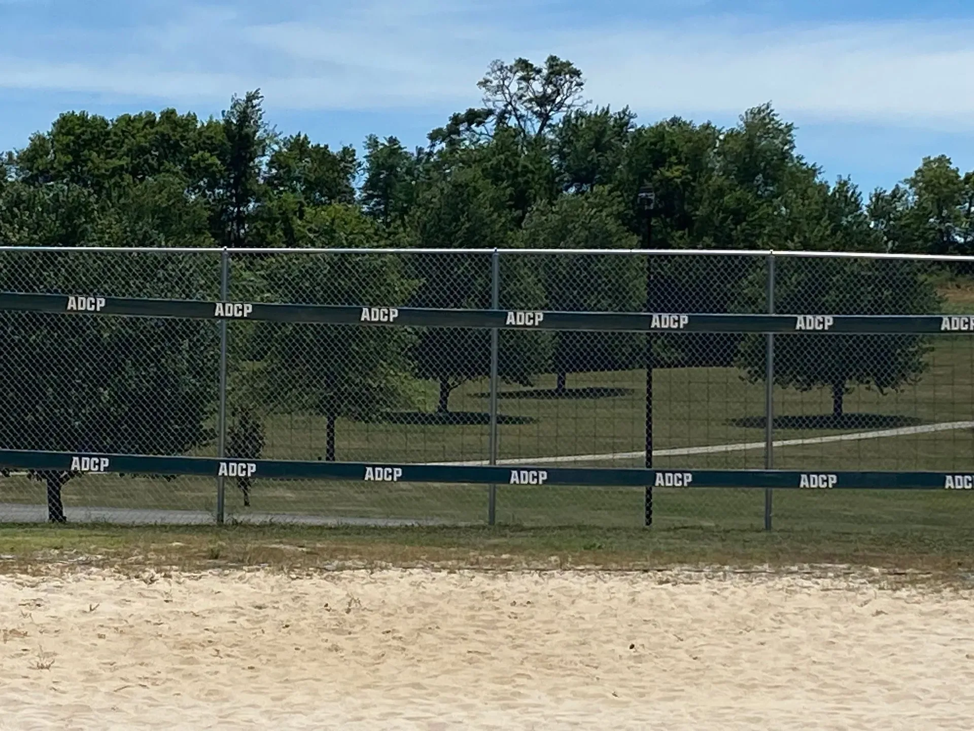 Chain-link fence with green trim in front of a grassy park area with trees and a blue sky.