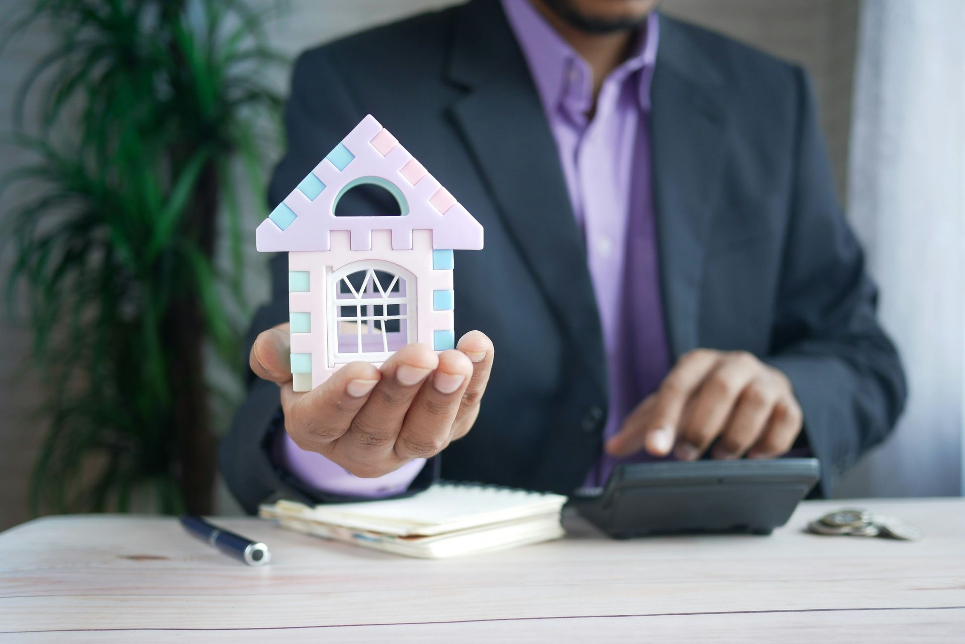 Man in suit holds toy house, uses calculator.
