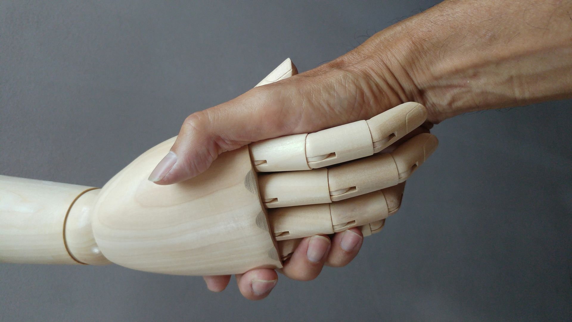 Human hand shaking a wooden mannequin hand against a gray background.