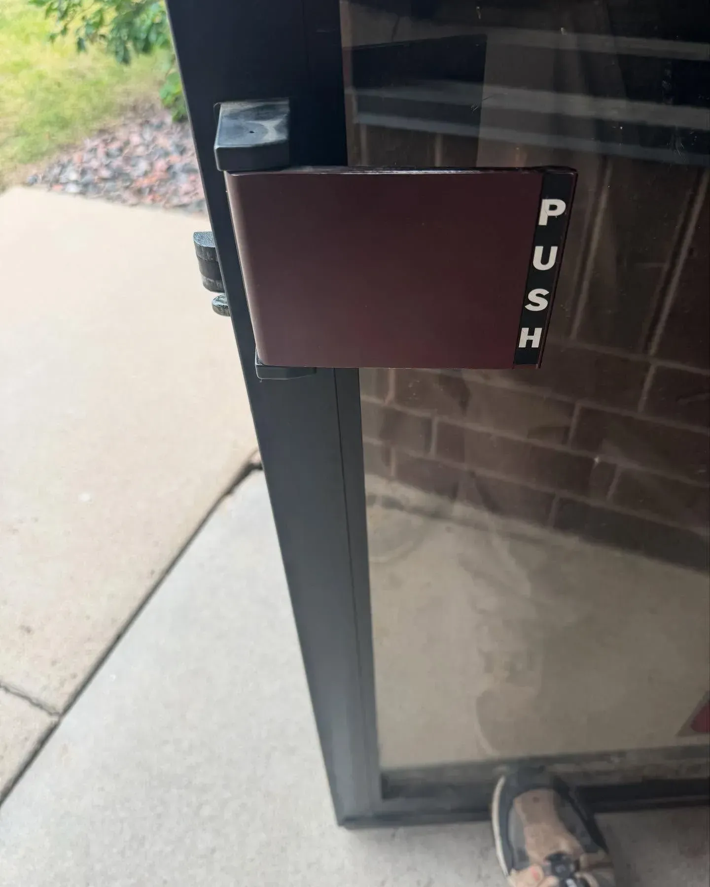 Glass door with a brown push plate and vertical “PUSH” label near the handle, concrete walkway outside