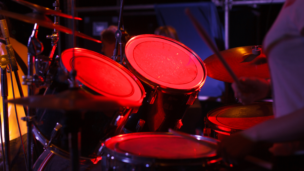A person is playing drums in a dark room with red lights behind them.