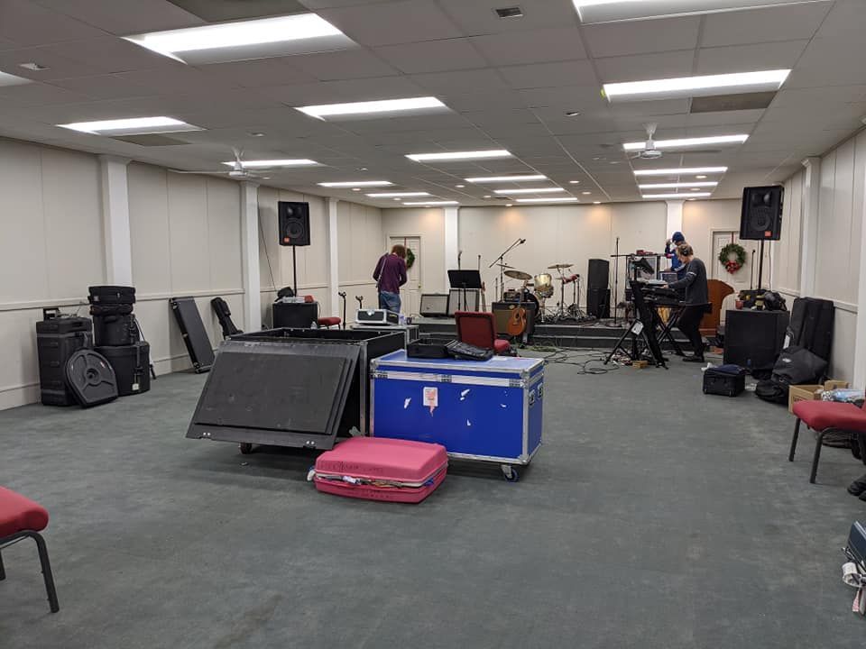 A band setting up equipment in a room. Speakers, drums, and instruments are visible, with people present.