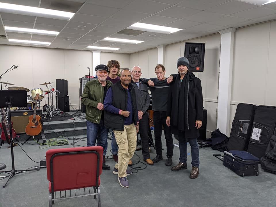 Group of musicians posing in a rehearsal space, with instruments in the background.