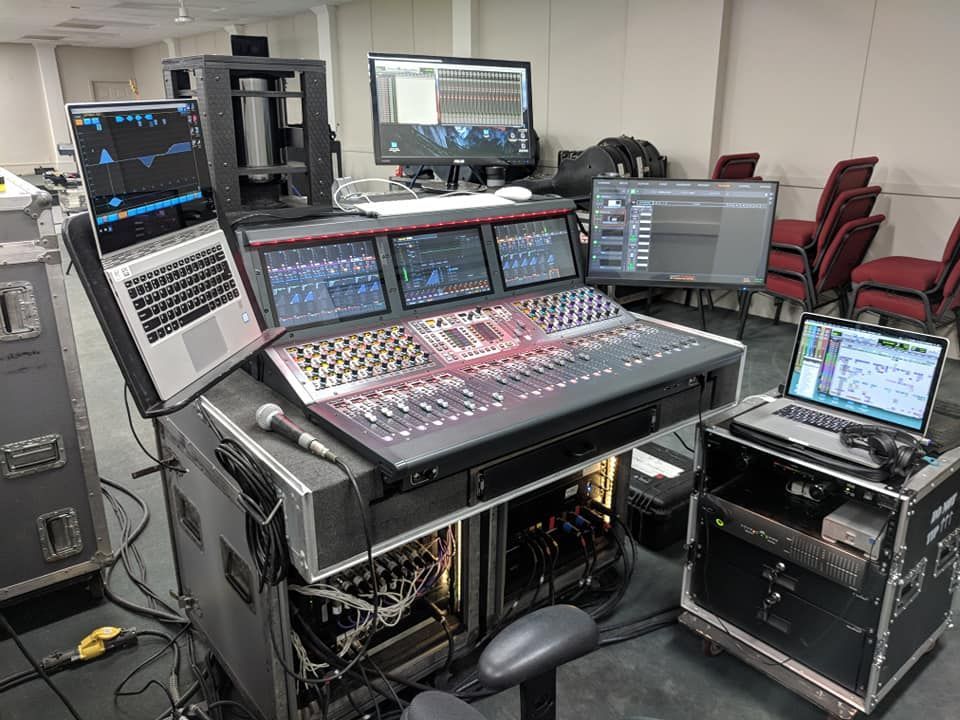 Audio mixing console setup with multiple screens and laptops, likely for a live event.