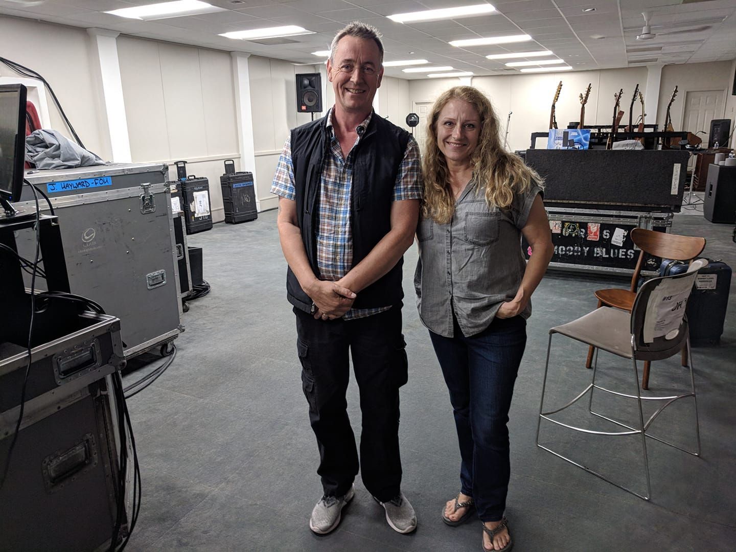 Two people pose inside a room with musical equipment. Man wears a vest and black pants.