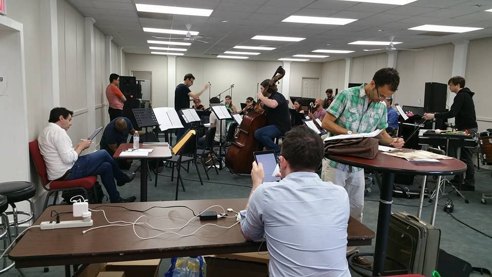 Musicians rehearse in a brightly lit room with instruments, music stands, and a double bass.