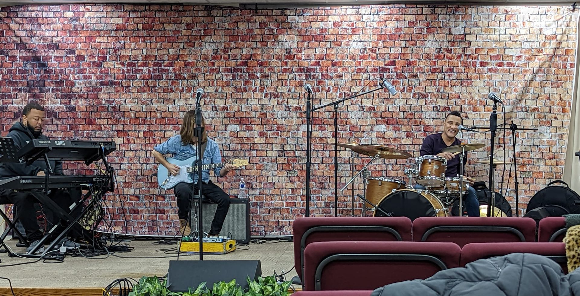 Band performing live on stage with a brick wall backdrop. Musicians playing guitar, drums, and keyboards.