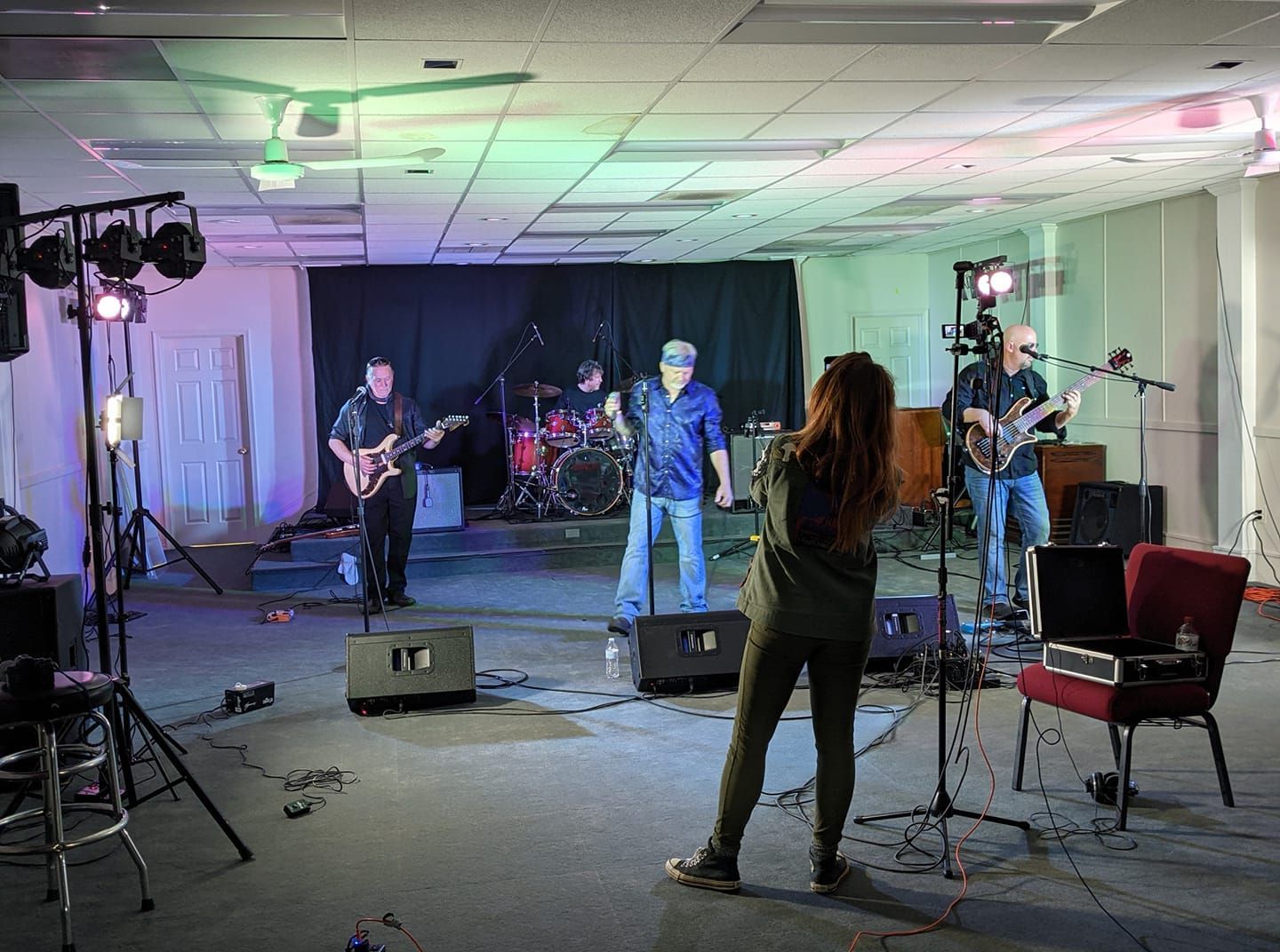 Band performing on stage, lit with colorful lights. Person filming them.