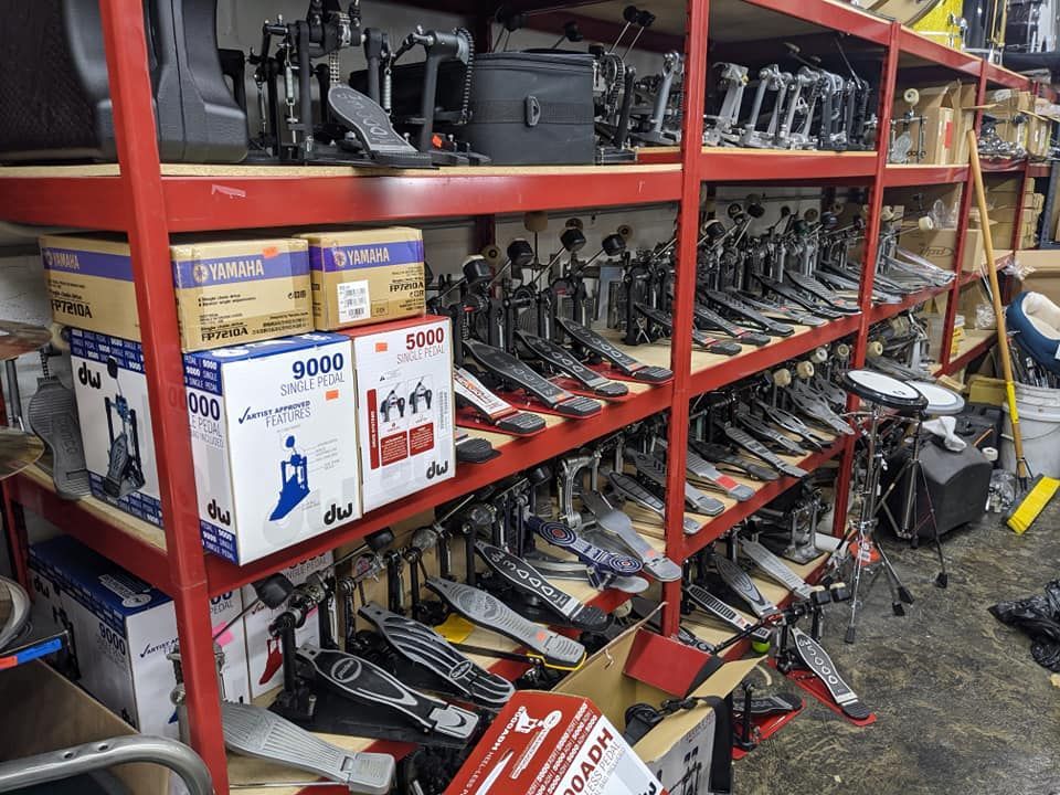 Shelves filled with drum pedals in a storage room. Boxes and other drum equipment are present.