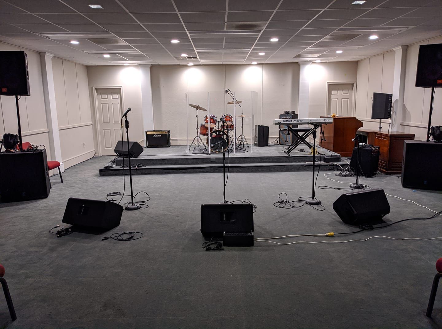 Acoustic equipment set up on a stage in a room, including a drum set, speakers, microphones, and a keyboard.