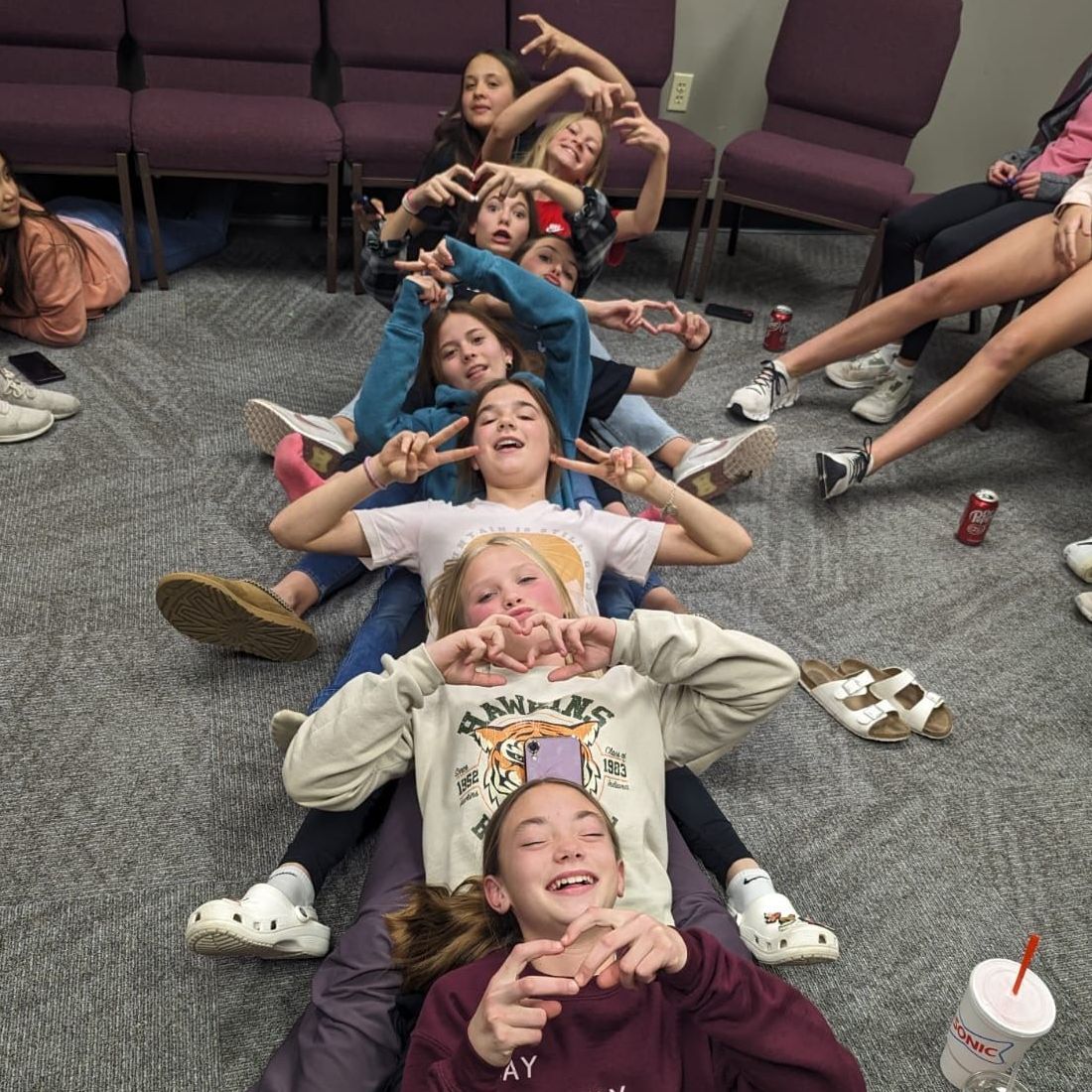 Group of girls posing in a line on the floor, making heart and peace signs. Indoors near chairs.