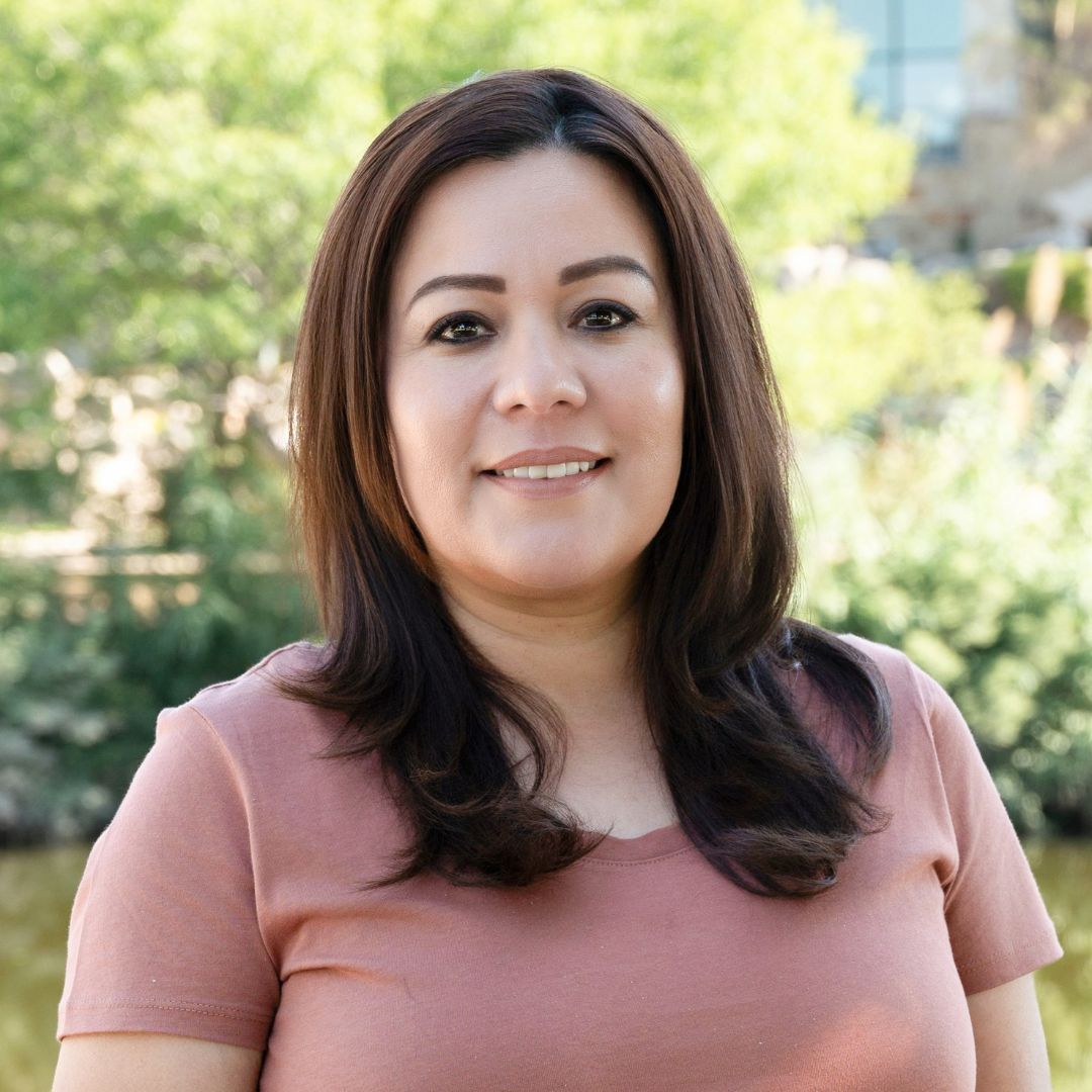 Man with a gray beard and blue shirt smiles outdoors. Brick building background with soft lighting.