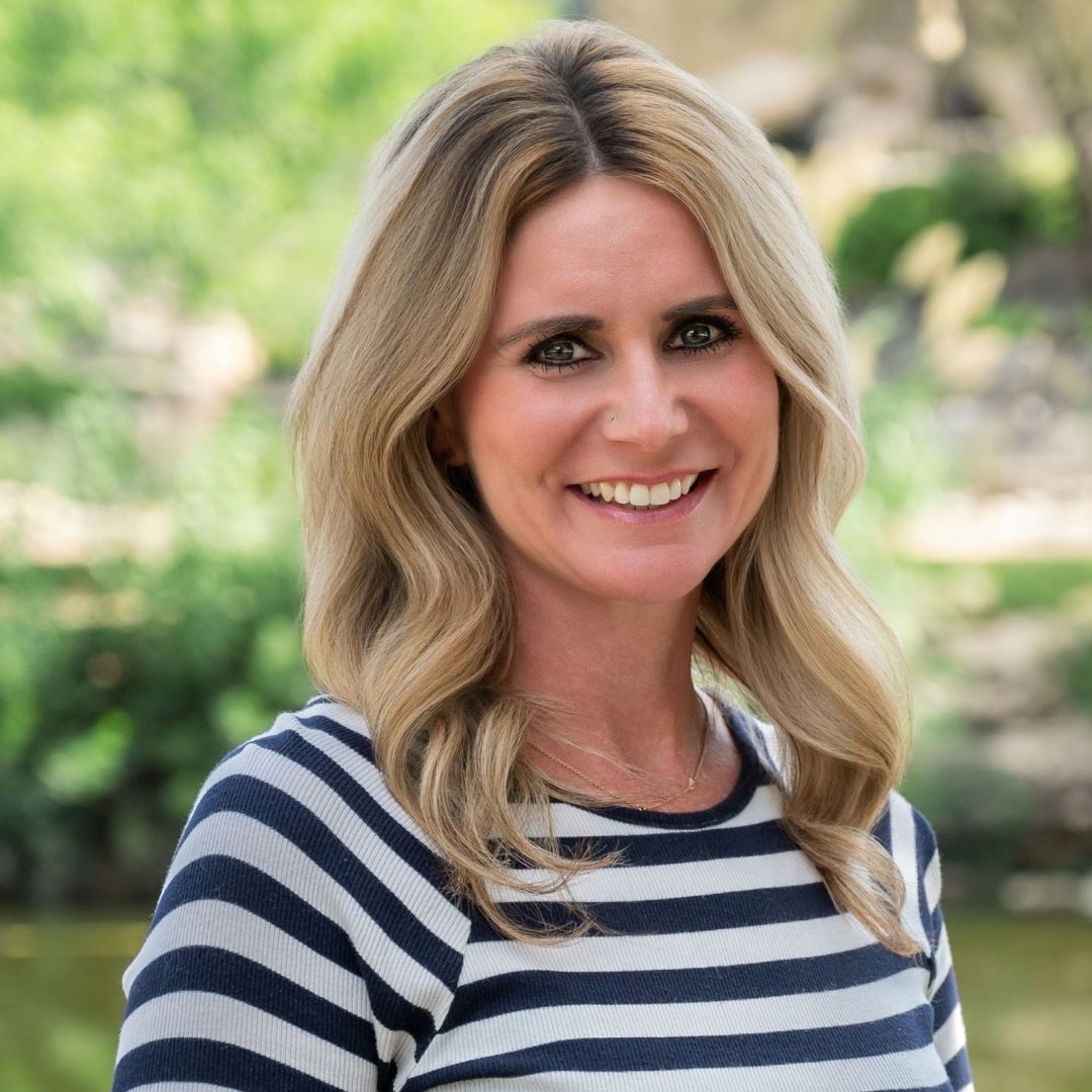 Bald man with glasses and beard, in a blue button-down shirt, smiles outdoors.